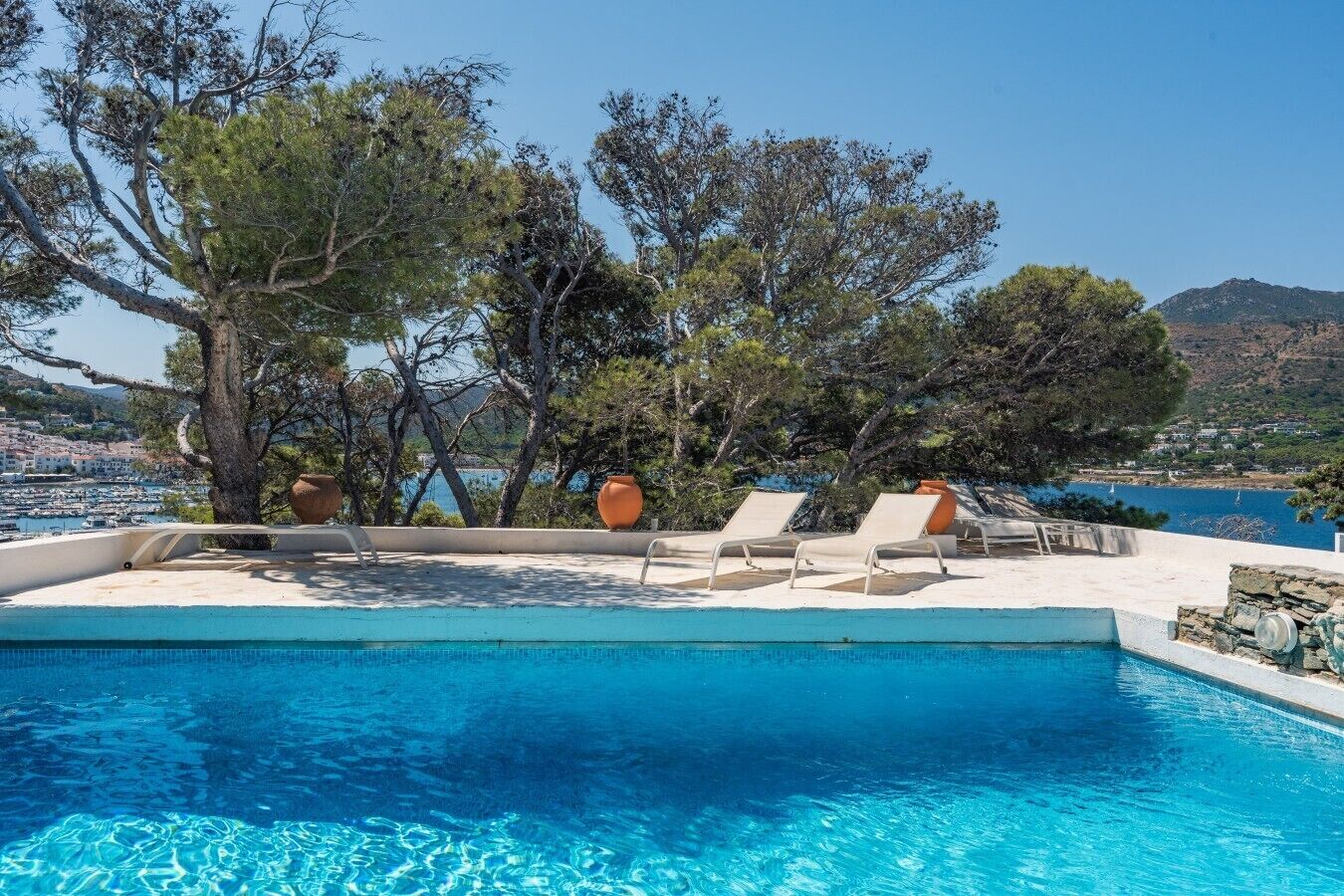 A pool with clear blue water, surrounded by lounge chairs and potted plants, overlooking a coastal town and hills.