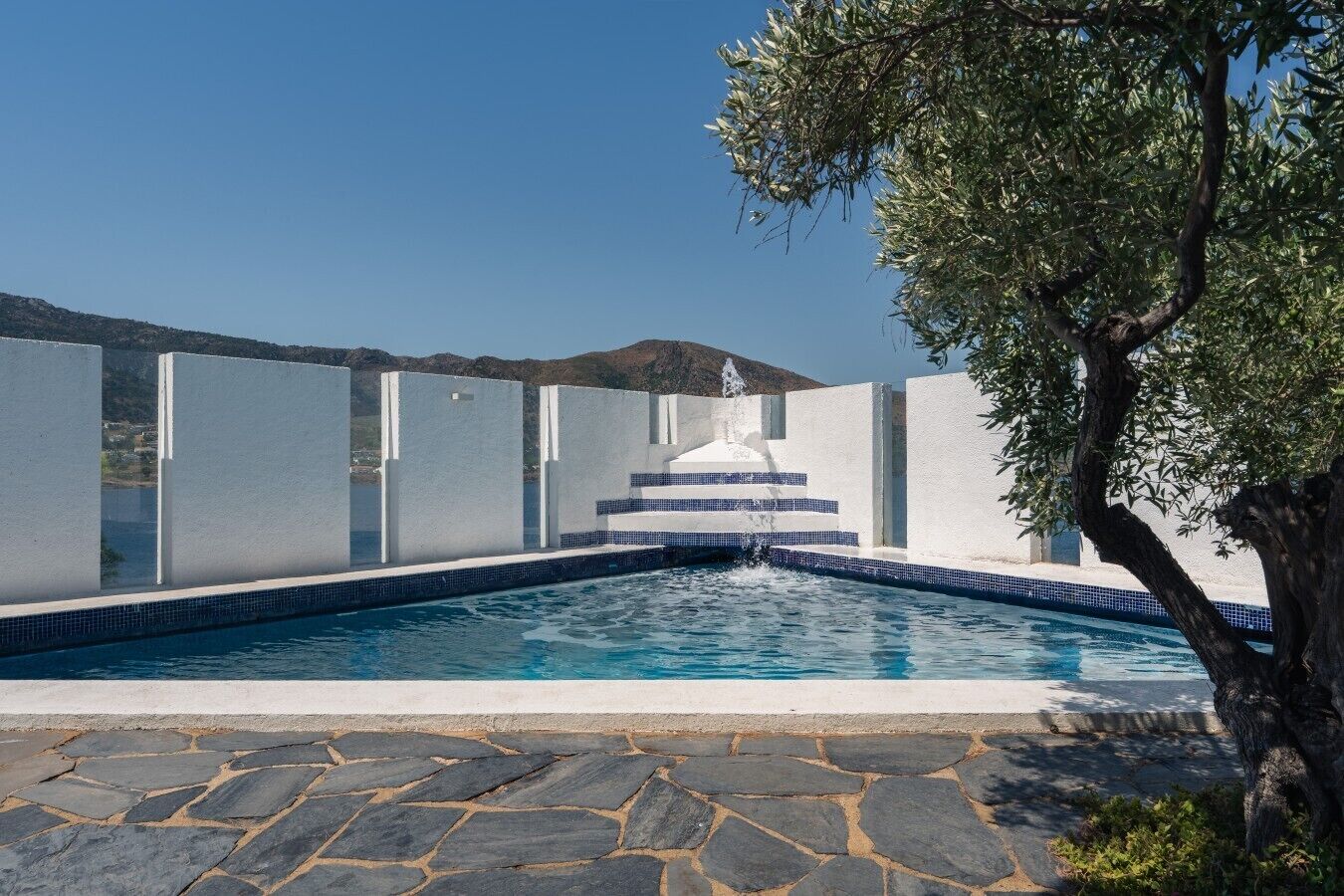 A swimming pool with a fountain, surrounded by white walls and stone flooring.