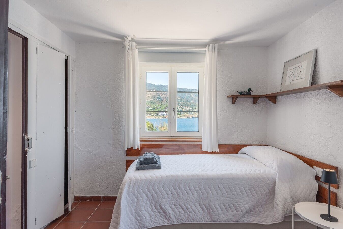 A bedroom with a bed, a bedside table, a window with curtains, and a view of the outdoors.
