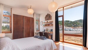 Un dormitori amb un llit gran, un armari de fusta i un escriptori amb cadira. Hi ha un gran finestral amb vista a una platja i edificis.