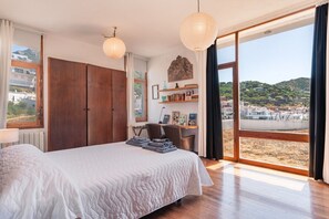 Ein Schlafzimmer mit einem großen Bett, einem Holzschrank und einem Schreibtisch mit Stuhl. Ein großes Fenster bietet Blick auf Strand und Gebäude.