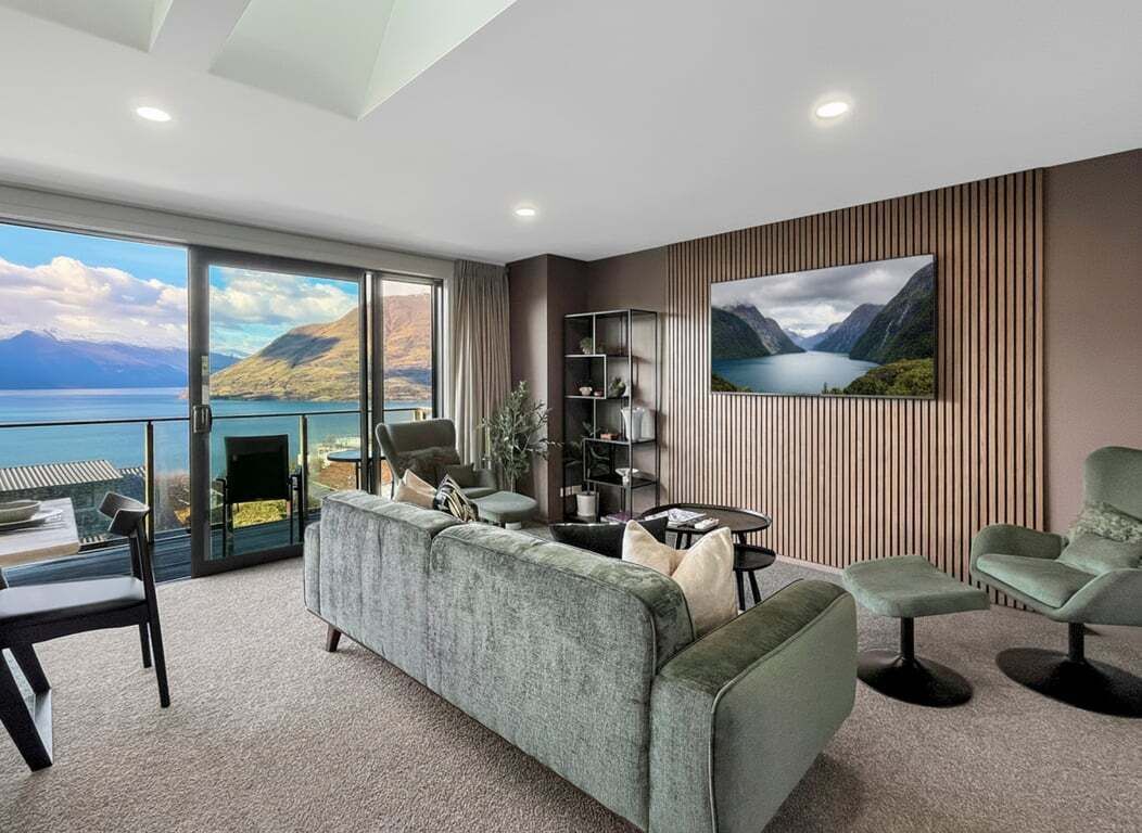 A modern living room with a large sofa, a coffee table, and a TV mounted on the wall. There is a bookshelf and a dining area with a table and chairs. The room has large windows offering a view of a lake and mountains.
