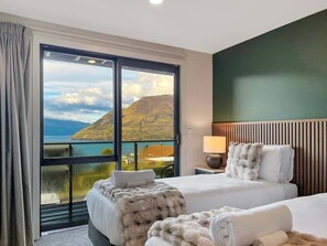 A hotel room with a large sliding glass door leading to a balcony with a scenic view of mountains and a body of water. There are two beds with white linens and fluffy blankets. - Queenstown 3-Bed Getaway | Alpine Views (Queenstown)