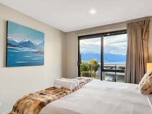 A hotel room with a bed, a fur throw, a painting of mountains, and a balcony with a view. - Queenstown 3-Bed Getaway | Alpine Views (Queenstown)