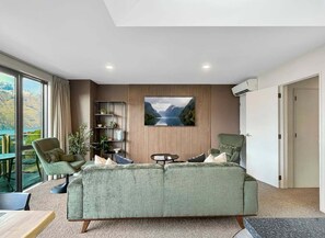 A modern living room with a green sofa, a coffee table, and a TV mounted on a wall with a scenic view. - Queenstown 3-Bed Getaway | Alpine Views (Queenstown)