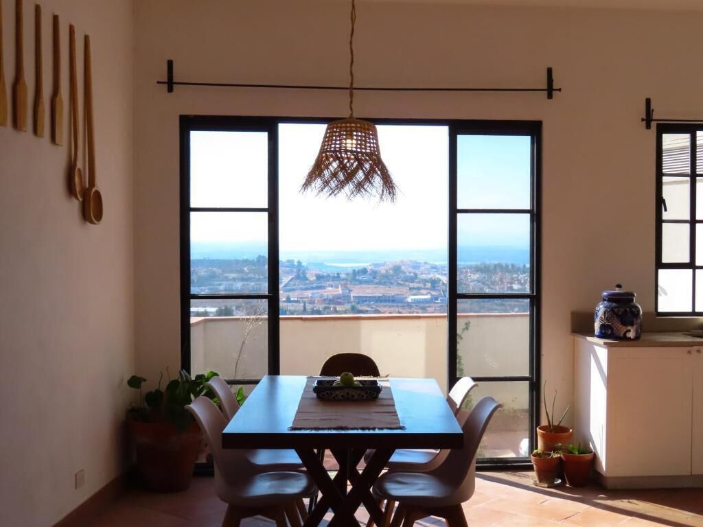 A dining area with a table and chairs, a hanging light fixture, and a view through large windows.