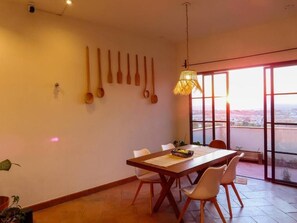 Uma área de refeições com mesa de madeira, cadeiras brancas e vista pela janela grande.