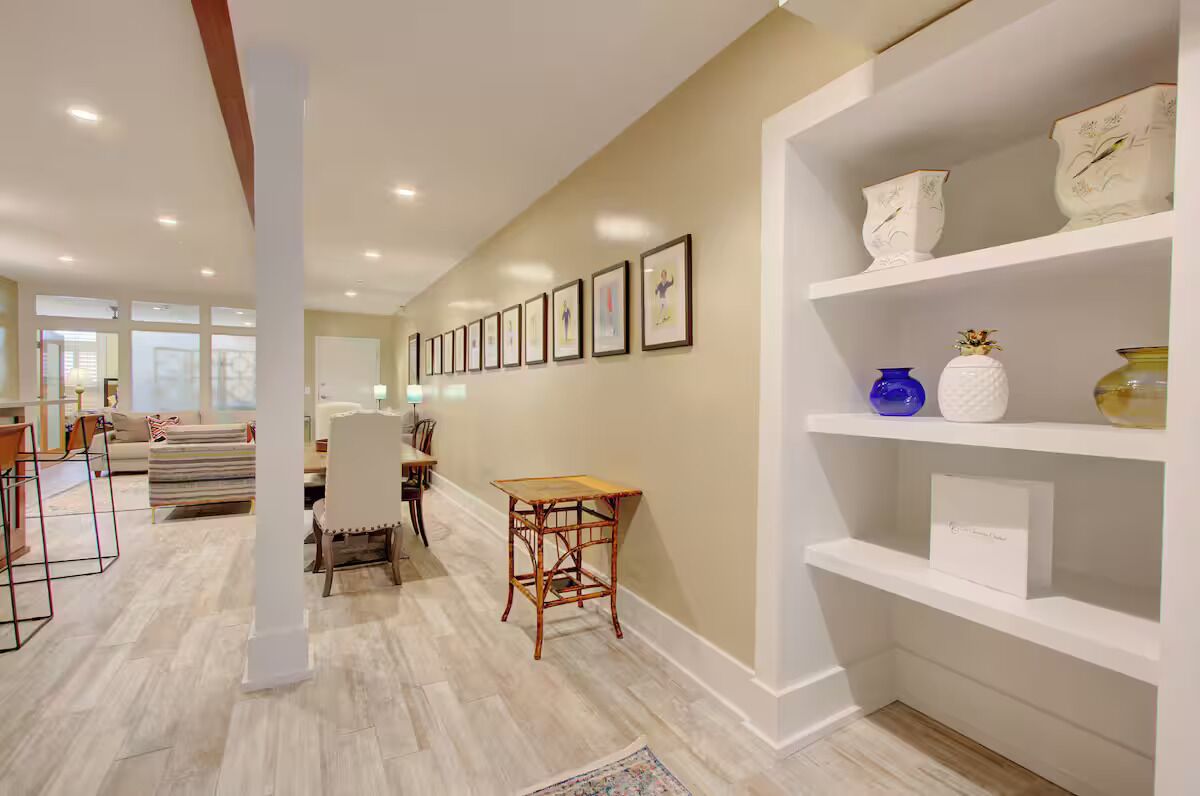 A spacious hallway with a dining area, framed artwork on the wall, and a white shelving unit with decorative items.