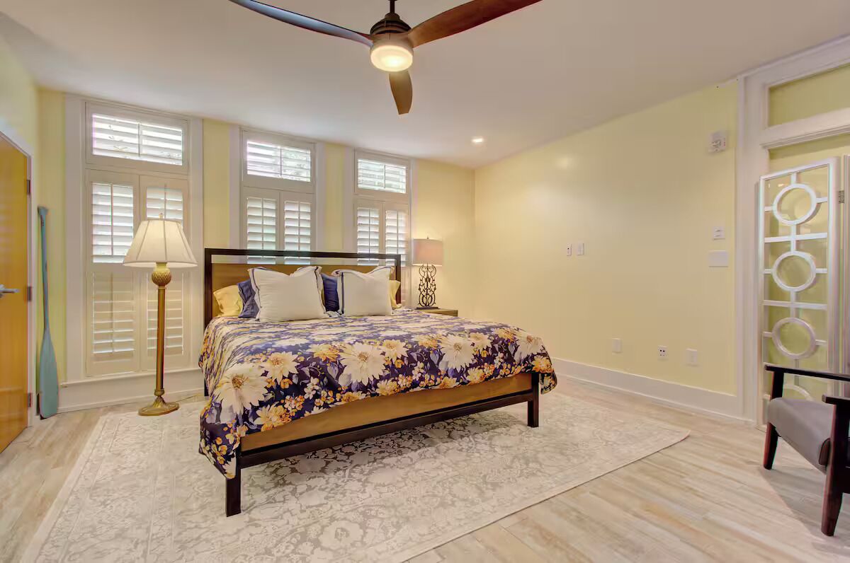 A bedroom with a bed, two bedside lamps, a ceiling fan, and a window with blinds.