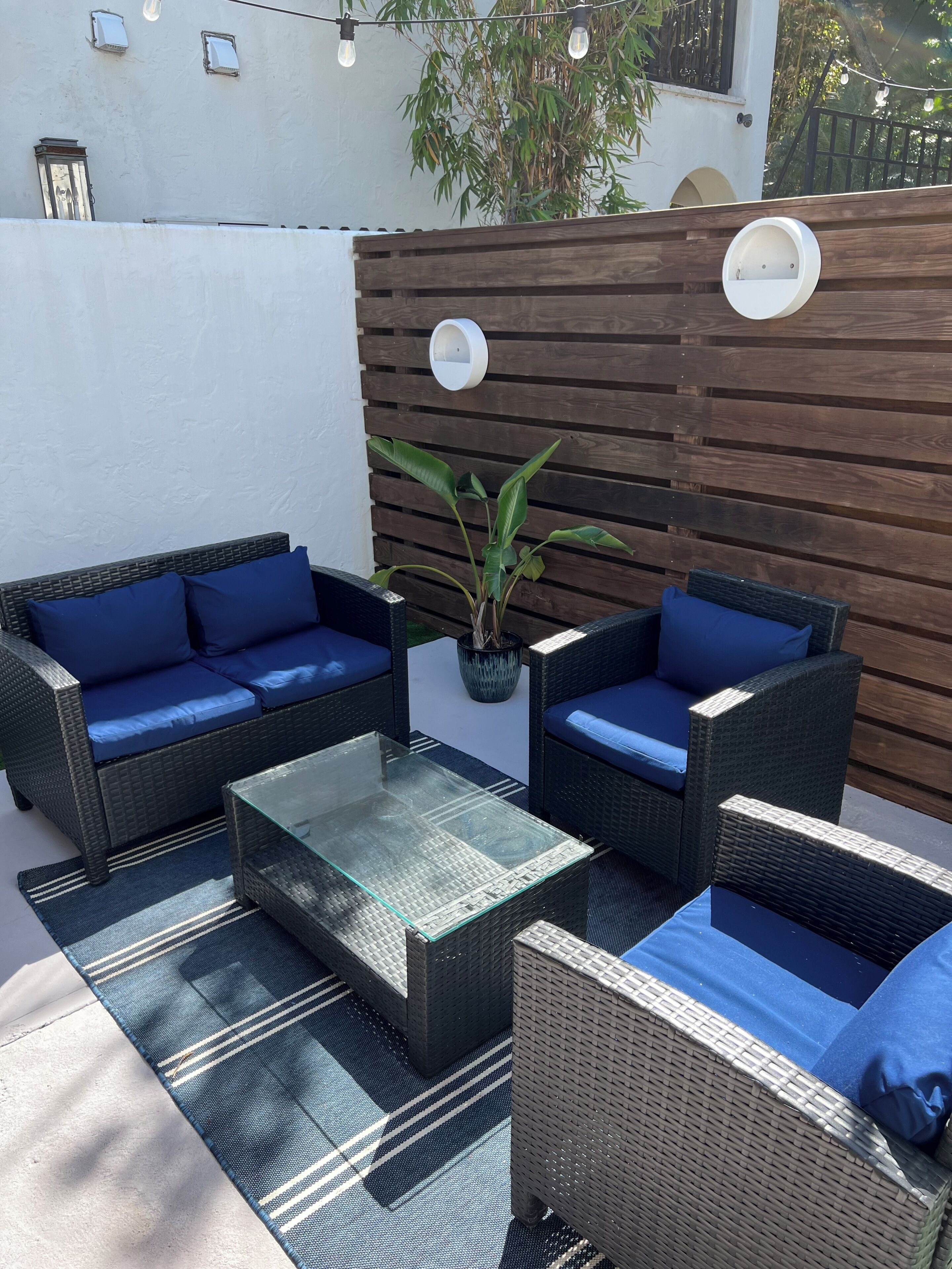 Un área de estar al aire libre con mobiliario de mimbre y cojines azules, una mesa de centro de vidrio y una planta en maceta.