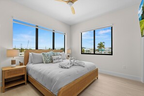 A bedroom with a bed, nightstands, a lamp, and two windows offering views of the outside.