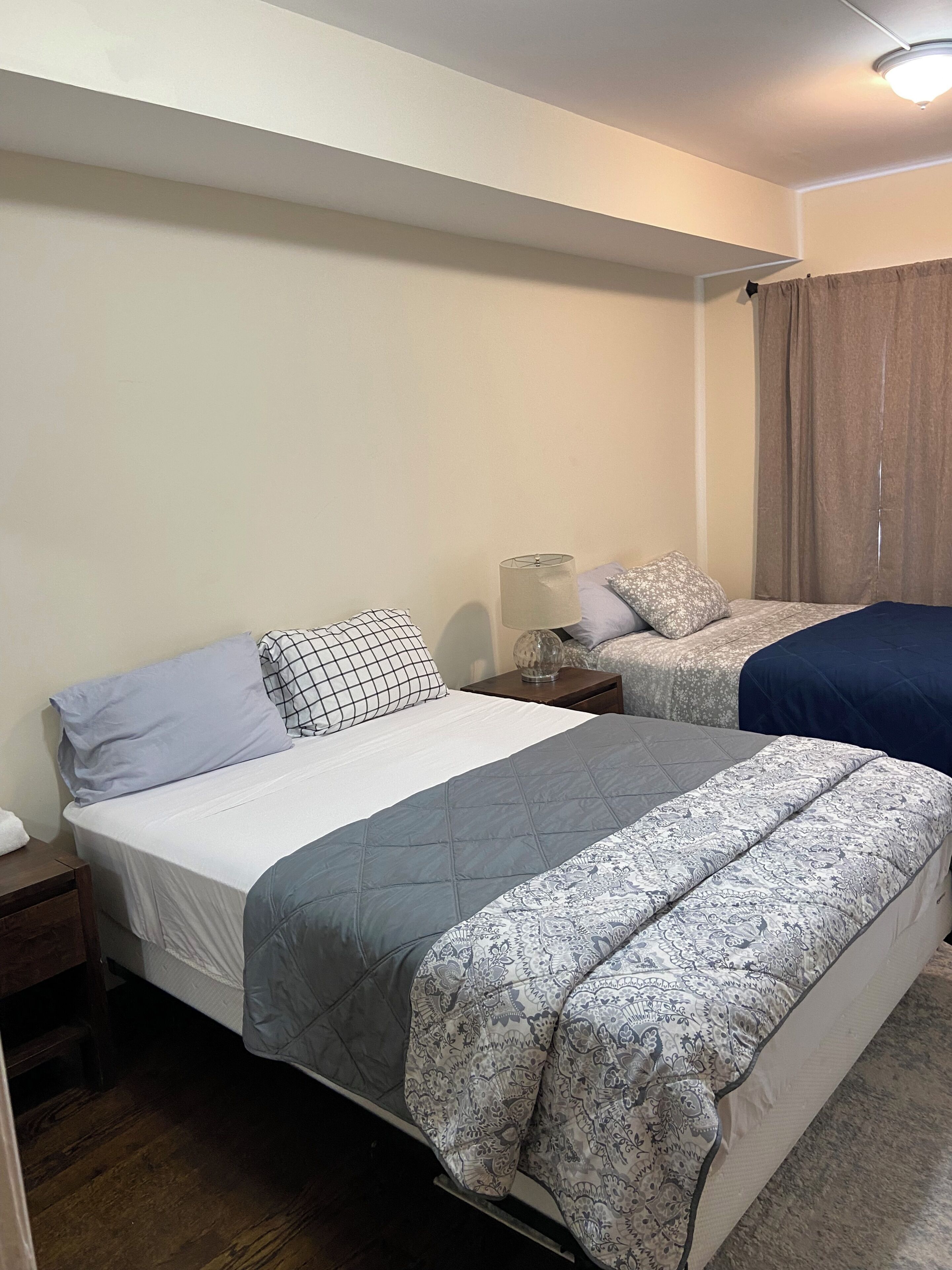 Quarto com duas camas, uma mesa de cabeceira, abajur e janela com cortinas.