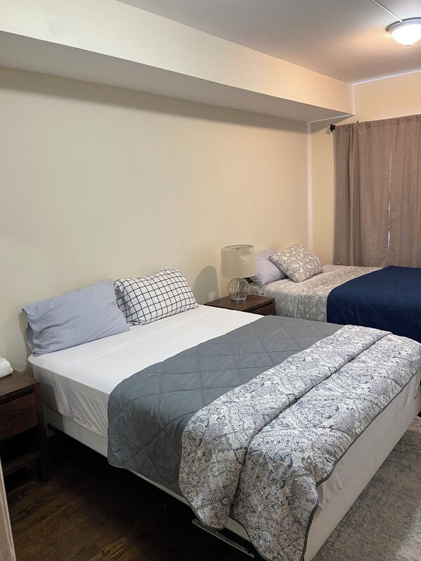 A bedroom with two beds, a nightstand, a lamp, and a window with curtains. - 6-bedroom apartment in terrific Jersey City with AC (Jersey City)
