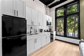 A modern kitchen with white cabinets, a black refrigerator, a stove, and a large window with a view of trees. - Explore Downtown Fun | Walk to Petco Park & More!! (San Diego)