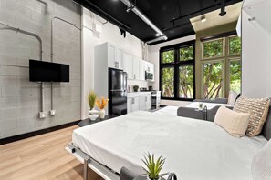 A modern bedroom with a large bed, a TV mounted on the wall, and a kitchen area with cabinets and appliances. - Explore Downtown Fun | Walk to Petco Park & More!! (San Diego)