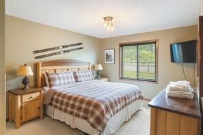 A bedroom with a bed, two bedside tables, a lamp, a television, and a window with blinds.