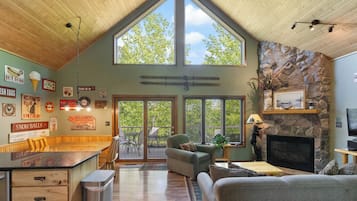 A spacious living area with a high ceiling, stone fireplace, and large windows overlooking a deck.