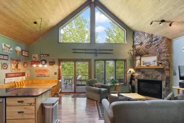 A spacious living area with a high ceiling, stone fireplace, and large windows overlooking a deck.