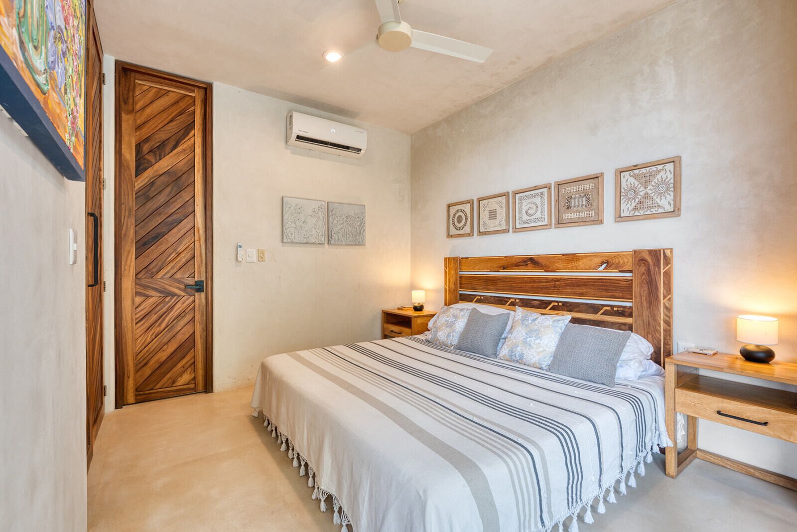 Ein Schlafzimmer mit Bett, Holz-Kopfteil, Nachttischen, Wand-Klimaanlage und Deckenventilator sowie einem gerahmten Kunstwerk.