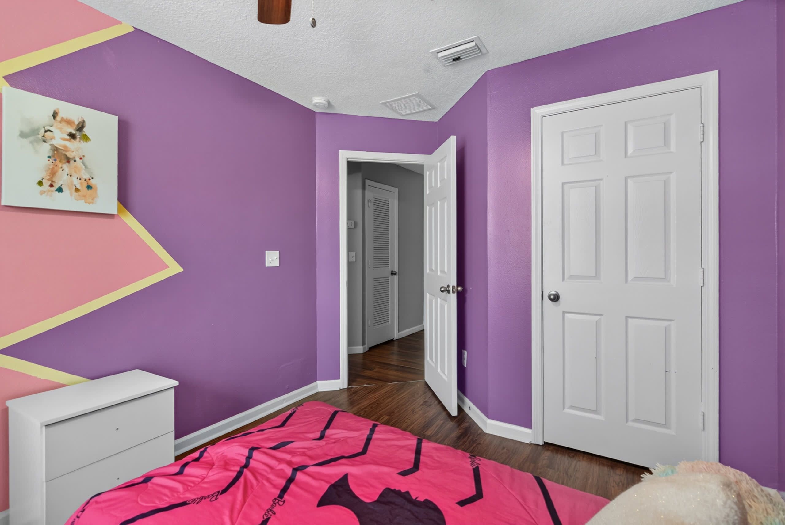 Une chambre avec des murs violets, une porte blanche, un lit recouvert d’une couvre-lit à motifs roses et noirs, et une commode blanche.