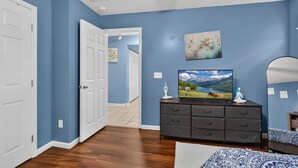 Una habitación con paredes azules, suelo de madera, una puerta blanca, un aparador con televisor y un cuadro.