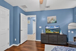 Ein Zimmer mit blauen Wänden, Holzboden, einer weißen Tür, einem Schminktisch mit Fernseher und einem Gemälde.