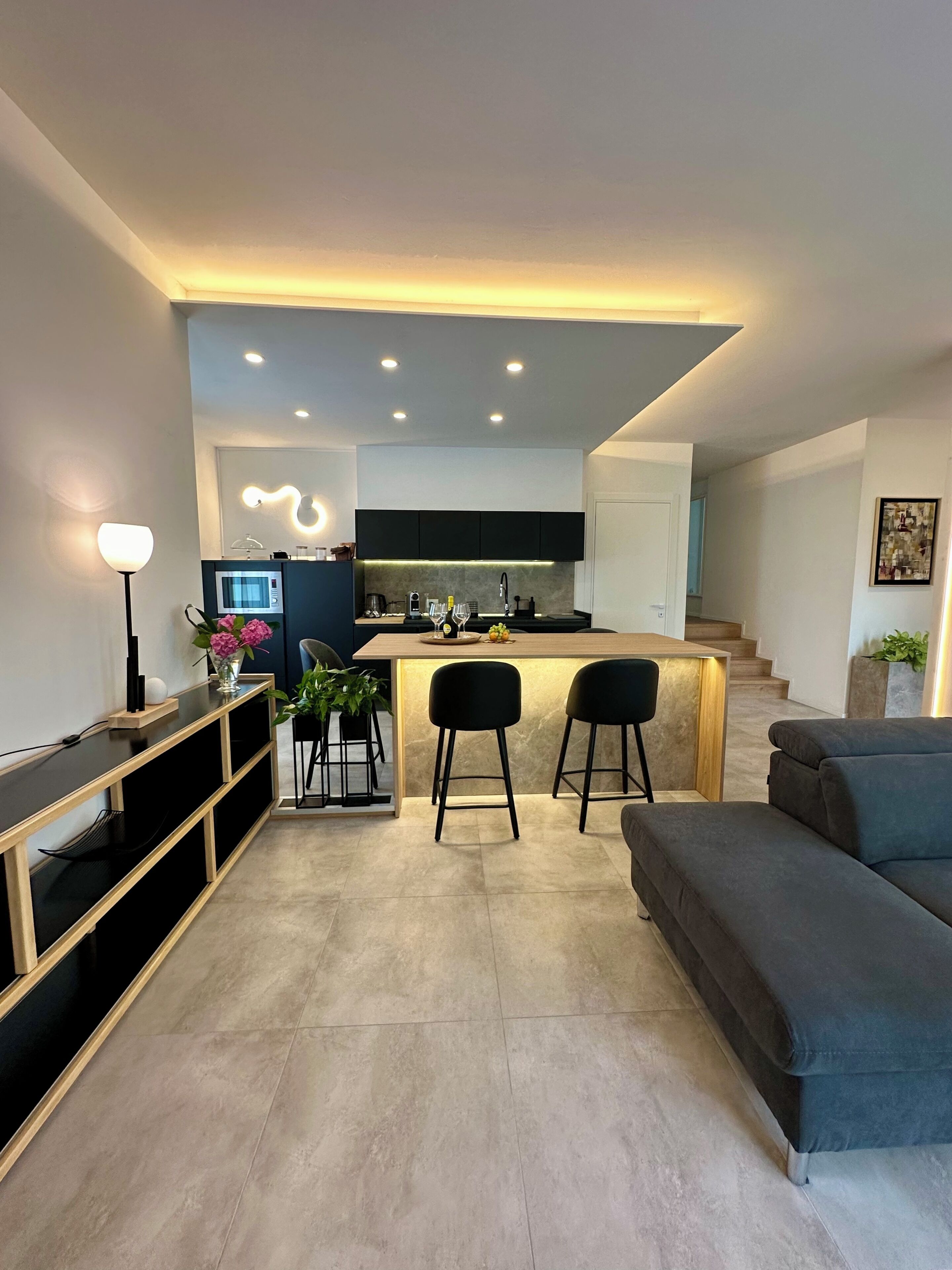 Una cucina moderna con isola centrale, sgabelli neri e un divano grigio in un ambiente soggiorno dalle linee contemporanee.