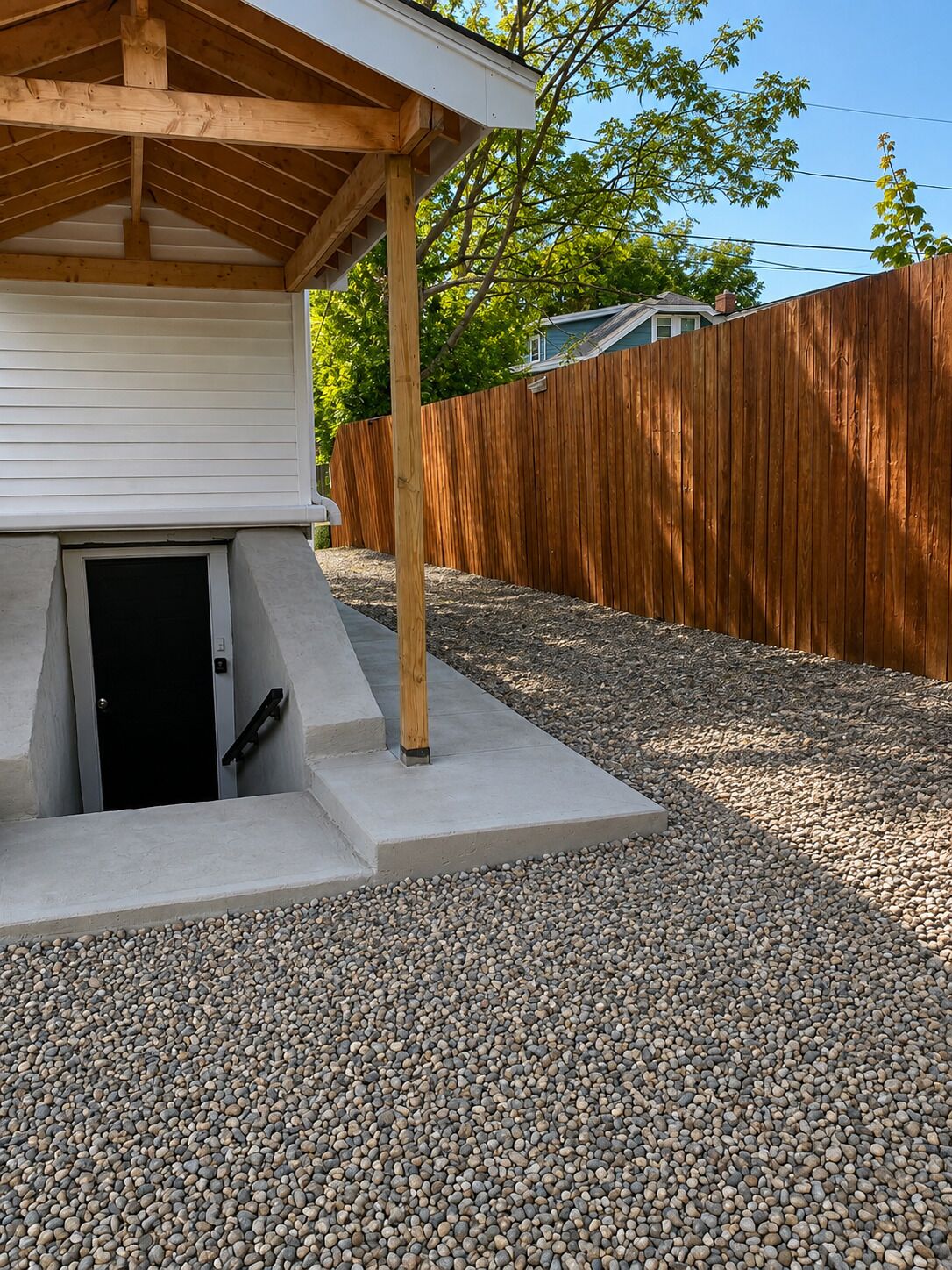 Un espace extérieur abrité, doté d’un sol en béton, d’un poteau en bois et d’un chemin de gravier menant à une porte.