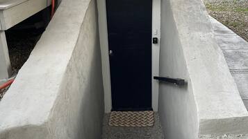 A concrete entryway leading to a dark door, with a wooden fence and vegetation in the background.