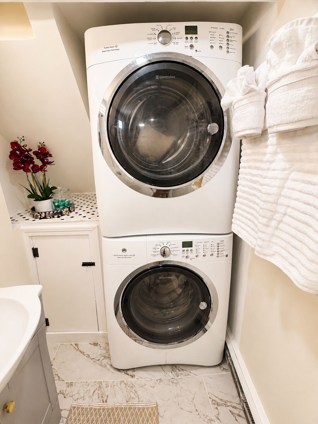 A compact, two-door front-loading washing machine in a bathroom with a marble floor, a small shelf with decorative items, and a towel rack.