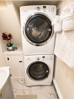 A compact, two-door front-loading washing machine in a bathroom with a marble floor, a small shelf with decorative items, and a towel rack.