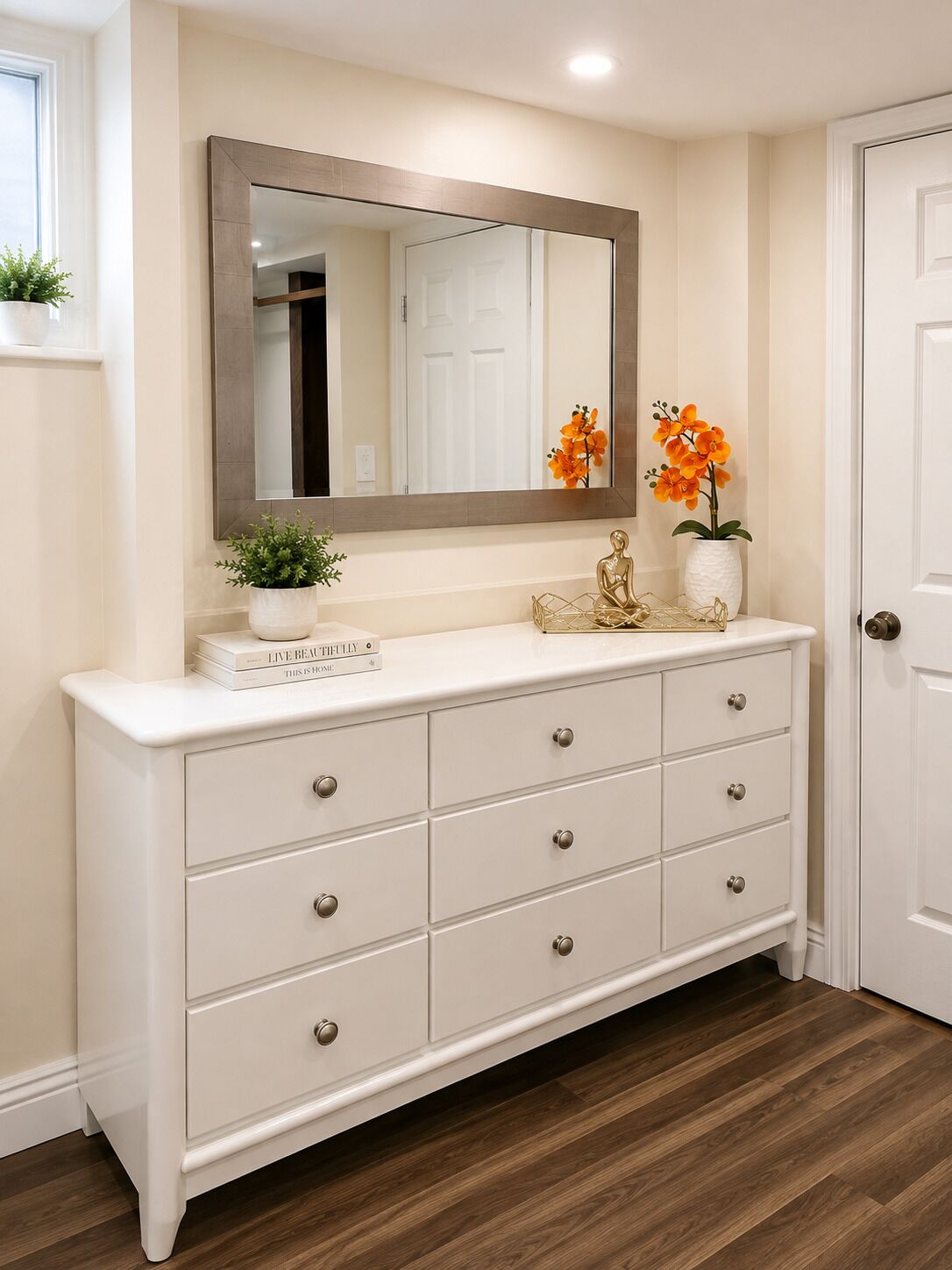A white dresser with six drawers, a large mirror above it, and two potted plants on the dresser.