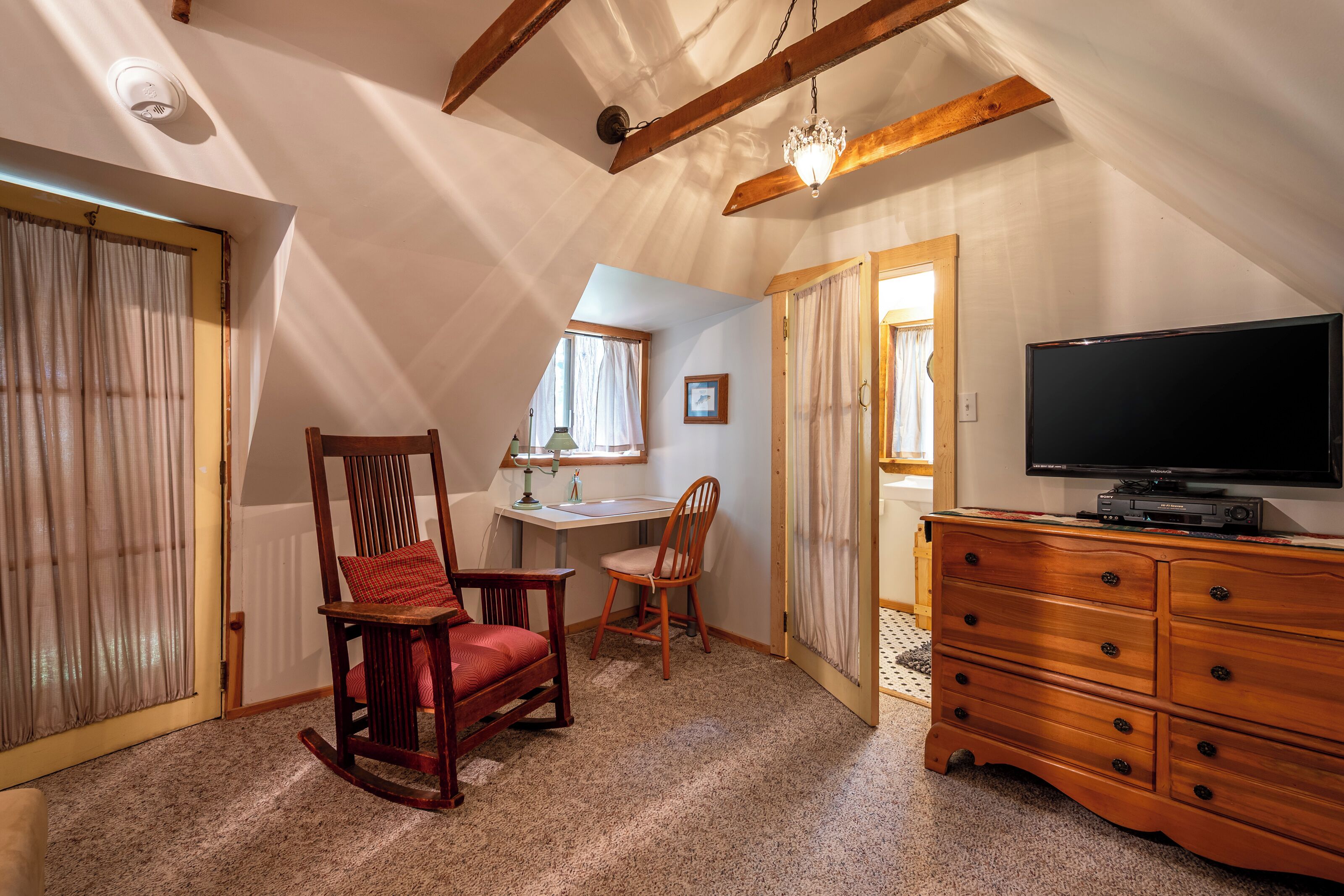 A cozy attic room with a sloped ceiling, a wooden rocking chair, a desk with a chair, a television, and a dresser.