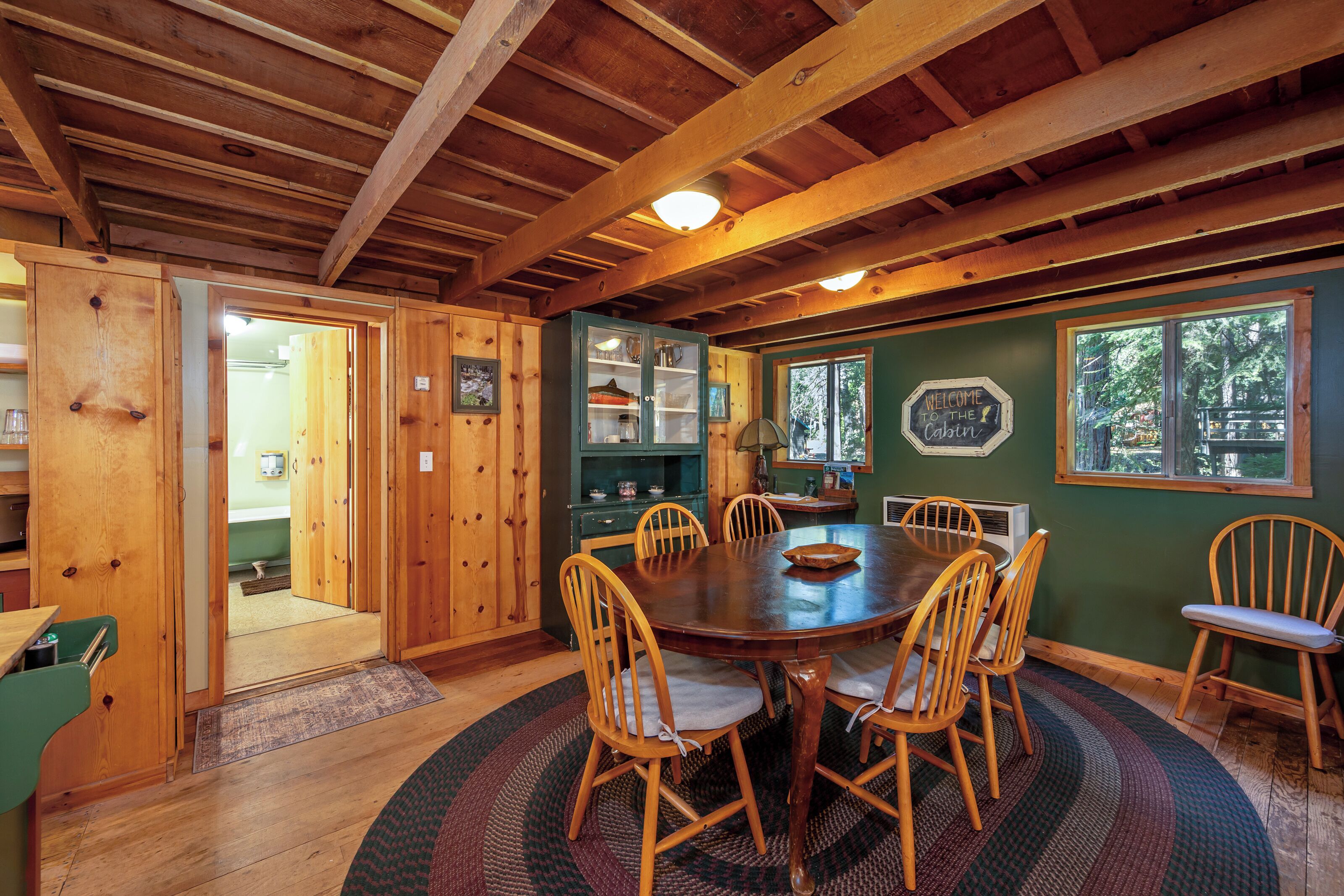 A wooden interior with a round dining table surrounded by chairs, a kitchen area with cabinets, and a view through a window.