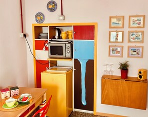 Eine kleine Küche mit Mikrowelle, Toaster und einem kleinen Kühlschrank. An den Wänden hängen Bilder in Rahmen, und ein Tisch ist mit einem Wasserkocher und Tassen bestückt.