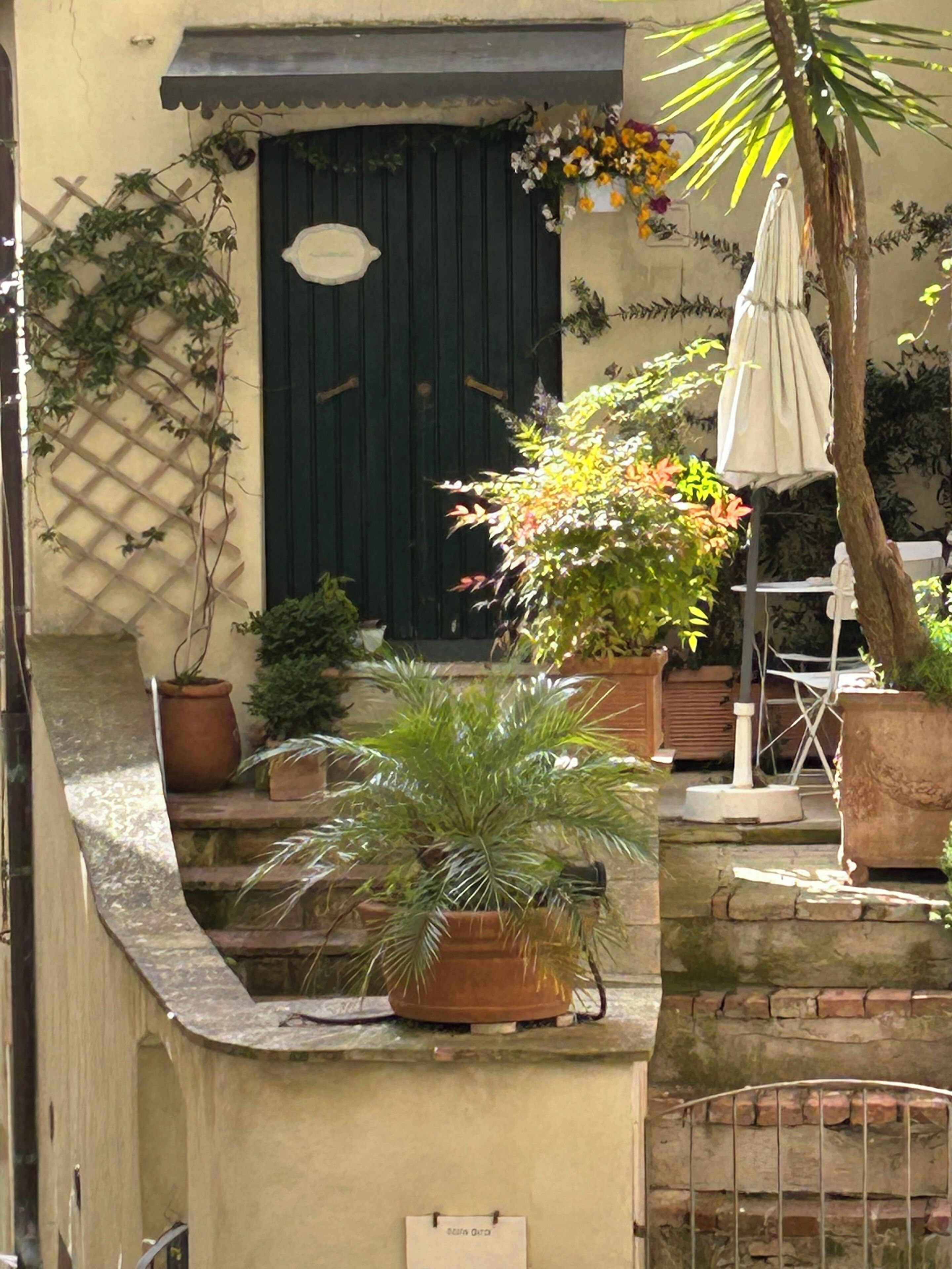 Un patio con un muro de piedra curvo, una puerta con placa decorativa, plantas en macetas y un paraguas.