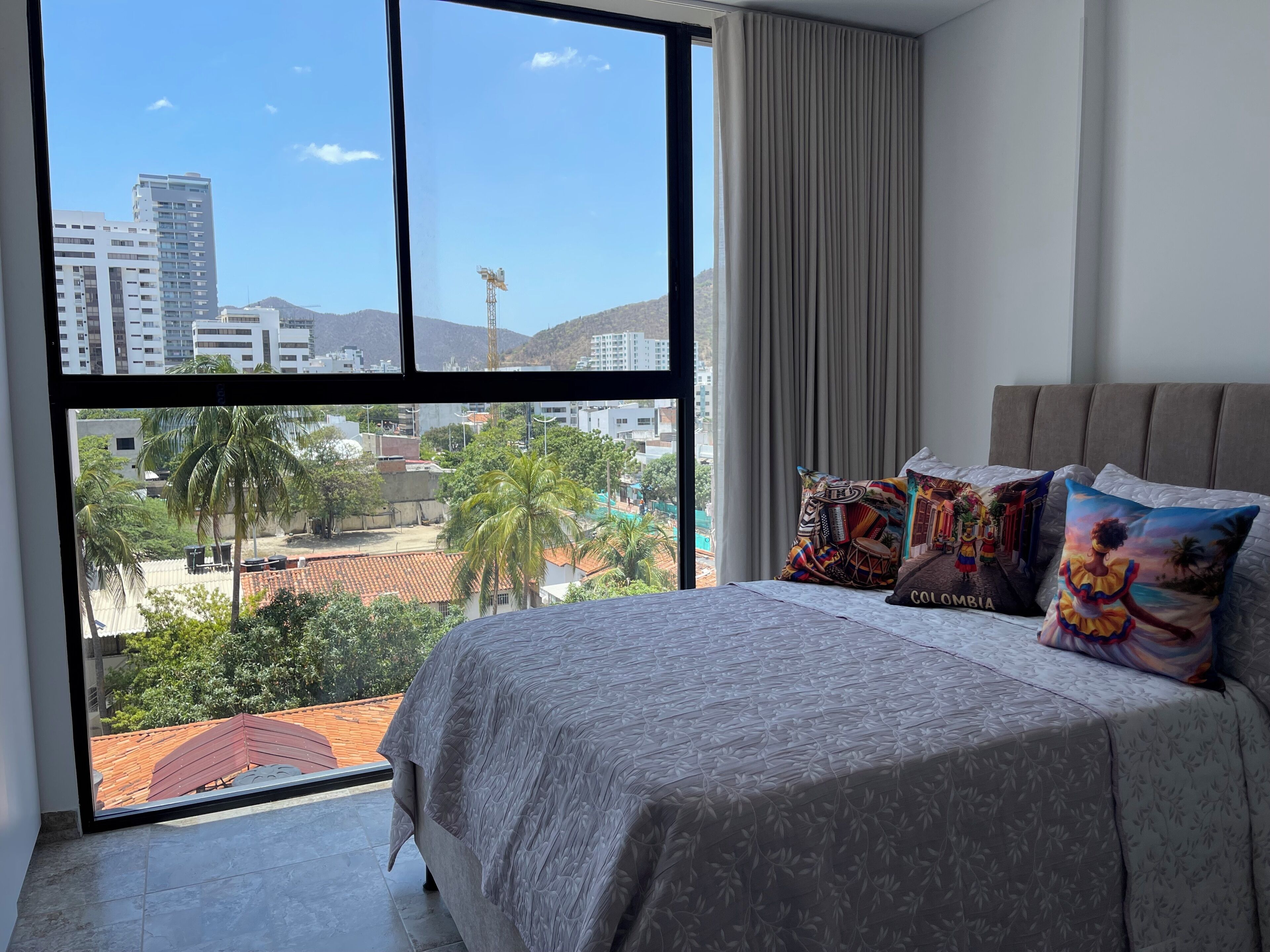 Un dormitorio con un ventanal grande, una cama con ropa de cama estampada y almohadones decorativos.