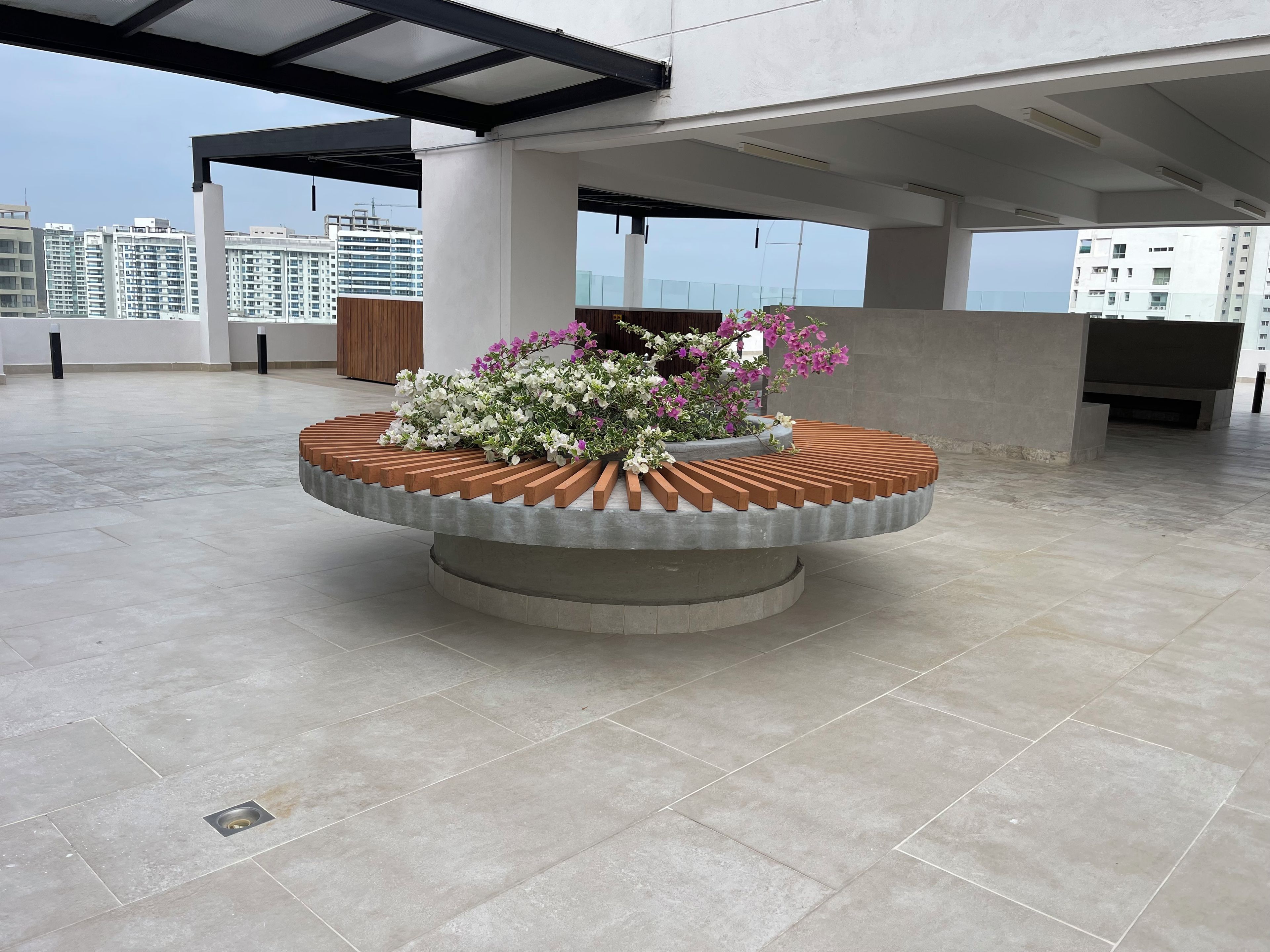 Eine Dachterrasse mit einem runden Blumenbeet, einer Holzbank und Blick auf hohe Gebäude.