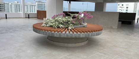 A rooftop terrace with a circular planter filled with flowers, a wooden bench, and a view of high-rise buildings.