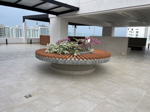 Terraço no telhado com um vaso circular cheio de flores, um banco de madeira e vista para edifícios altos.