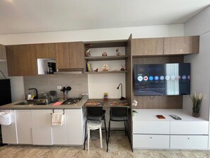 Eine moderne Küche mit Flachbildfernseher, der einen Homescreen anzeigt, einem Gasherd, einer Kaffeemaschine und einer kleinen Essecke mit zwei Stühlen.