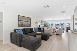 A modern living room with a sectional sofa, ottoman, and a view of the sea through large windows. - Coastal Reserve Executive Beach Home w/ golf cart (Indian Rocks Beach)