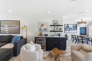 A modern living room with a sofa, armchair, and a coffee table. There is a kitchen area with a wine cooler and a dining table with chairs.