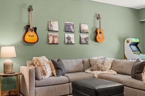 Ein Wohnzimmer mit einem Ecksofa, zwei Gitarren, einer Lampe und einer Spielkonsole.