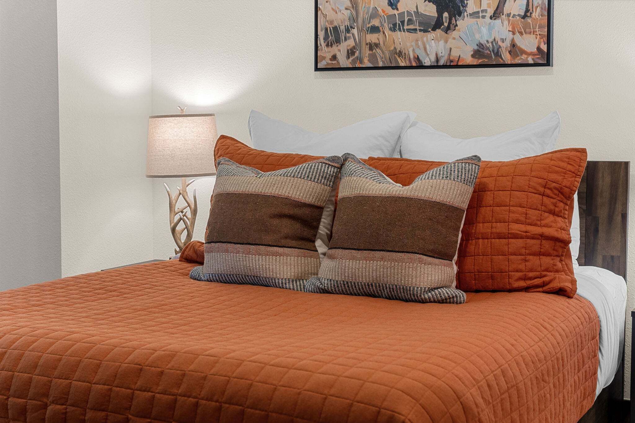 A bed with a quilted orange blanket, two patterned pillows, a white pillow, and a wall-mounted painting.