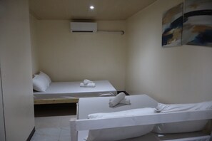 A small, single-room hotel room with two beds, a ceiling-mounted air conditioning unit, and a painting on the wall.