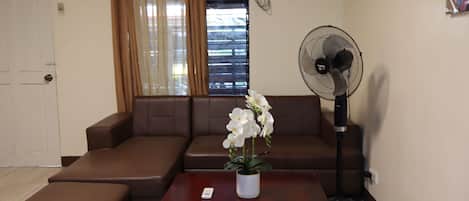 Una sala de estar con un sofá marrón, una mesa de centro de madera con un control remoto, un florero blanco y un ventilador de pie.