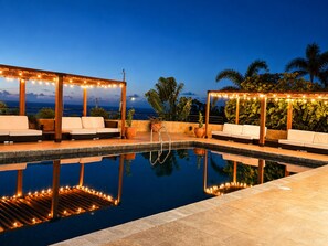 A poolside area with lounge chairs and string lights, surrounded by palm trees and a view of the ocean. - Luxury 5-bed, 5-bath with AC, pool, beach, hot tub, butler, chef, golf, sea view (Montego Bay)