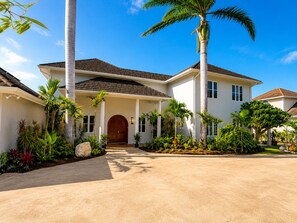 A two-story house with a central entrance, surrounded by a variety of tropical plants and palm trees. - Luxury 5-bed, 5-bath with AC, pool, beach, hot tub, butler, chef, golf, sea view (Montego Bay)