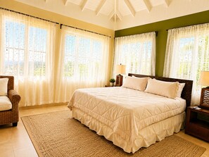 A bedroom with a large bed, two bedside tables, a chair, and a window with curtains. - Luxury 5-bed, 5-bath with AC, pool, beach, hot tub, butler, chef, golf, sea view (Montego Bay)